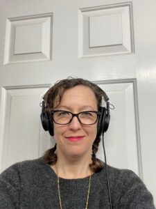 A picture of Isabelle with her brown hair in braids. She is wearing over the ear headphones, black glasses and red lipstick. She is smiling in a mischievous way.