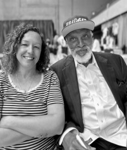 A black and white picture of Isabelle with john a powell in a conference setting. Isabelle is a white woman with mid-length brown curly hair and a striped t-shirt. john is a Black man with a white beard. He is wearing a white dress shirt with a black suit jacket and a black baseball hat that says Bridger in white letters.